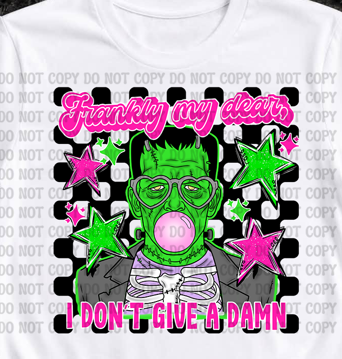 A white short-sleeve graphic T-shirt laid flat on a dark Halloween-themed background with skulls, pumpkins, and skeleton props. The shirt features a colorful front print with pink text reading 'Frankly my deers I DON'T GIVE A DAMN' and a green monster face surrounded by stars and puzzle-piece shapes.
